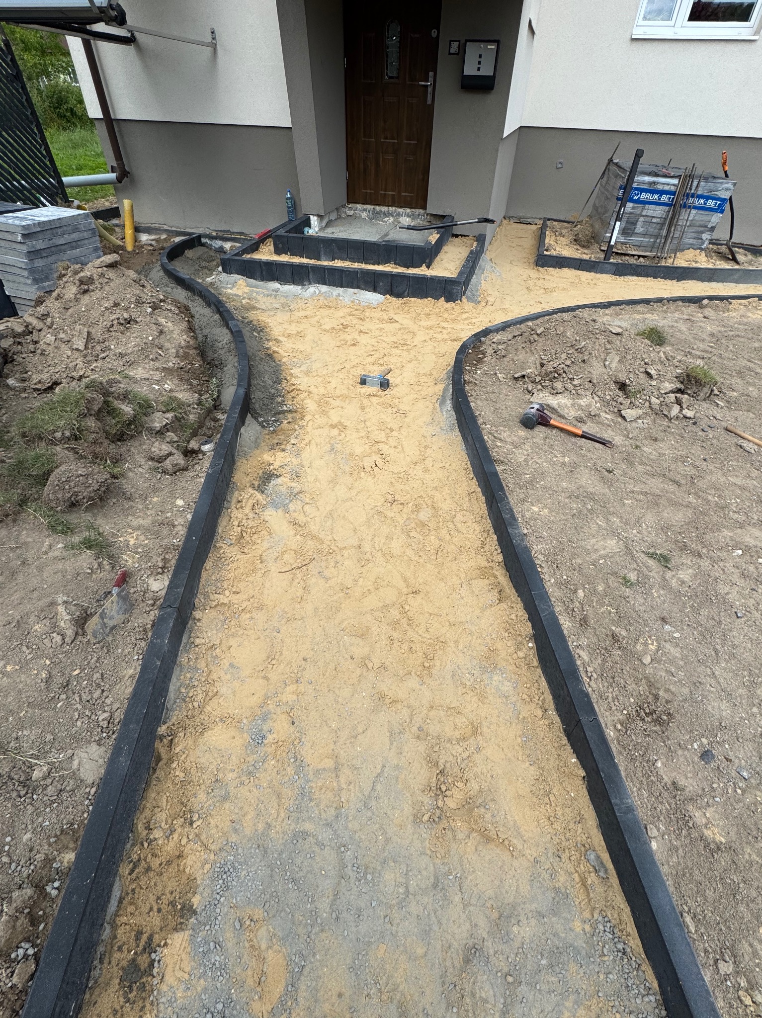 Przygotowanie podłoża pod ścieżkę z kostki brukowej: obrzeża ułożone, piasek rozsypany, widoczne narzędzia i zapas kostki. Etap prac przed ułożeniem nawierzchni.