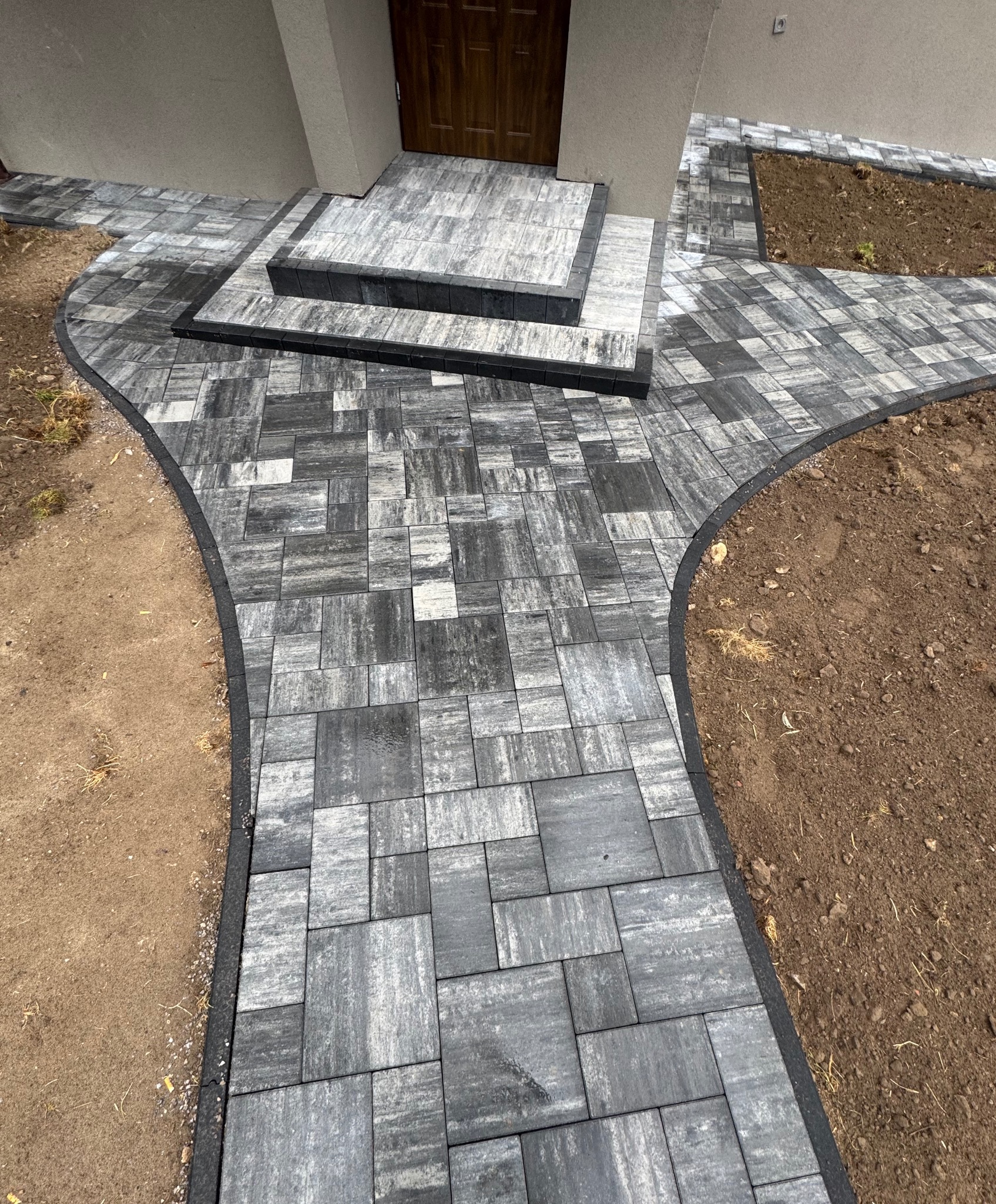 Ścieżka z kostki brukowej w odcieniach szarości prowadząca do wejścia domu, obrzeża wykończone czarnym elementem. Fragment rabaty z ziemią po prawej stronie.