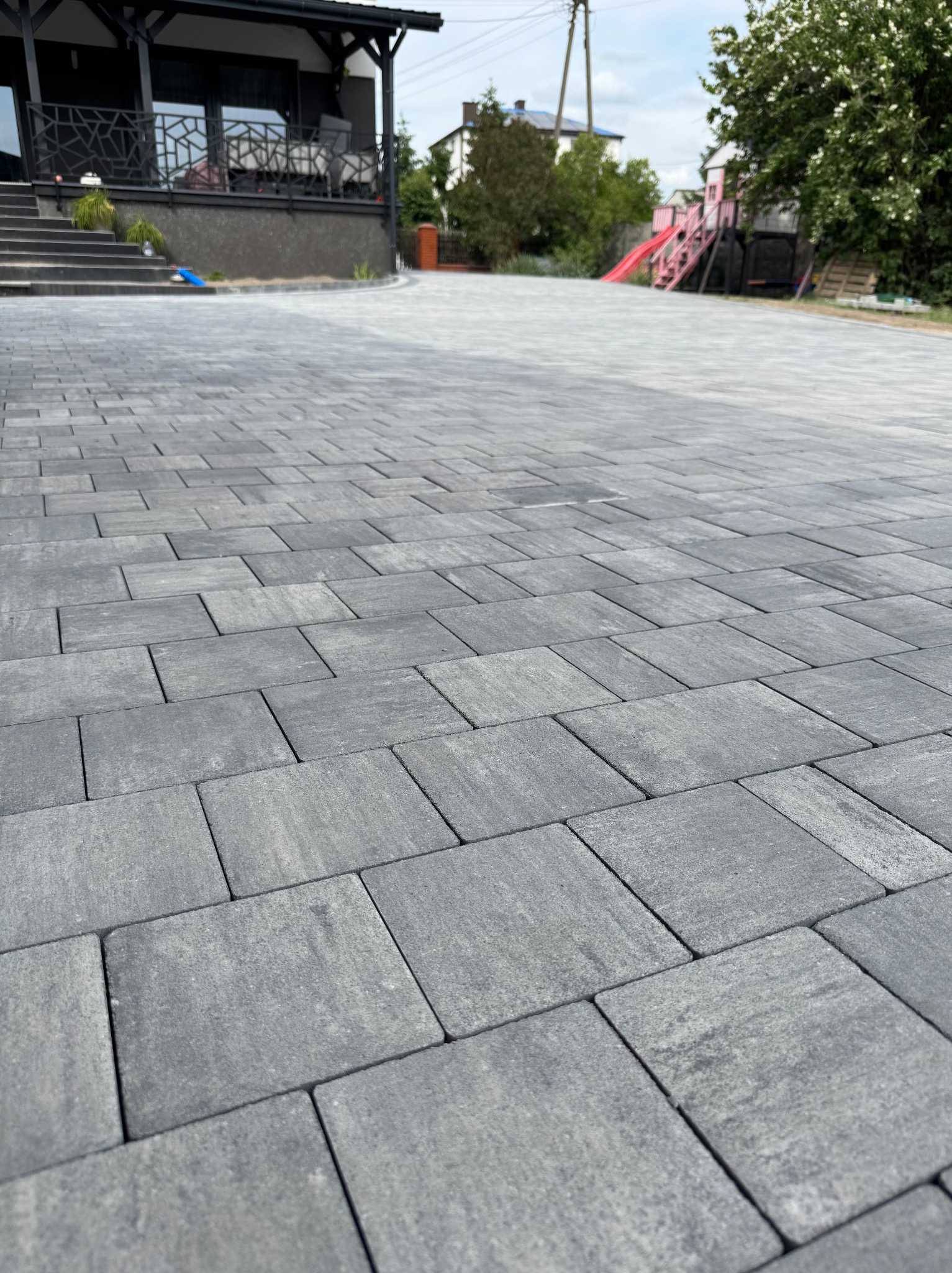 Ułożona szara kostka brukowa prowadząca do domu z tarasem. W tle widoczny plac zabaw dla dzieci. Perspektywa z dołu podkreśla regularność wzoru.