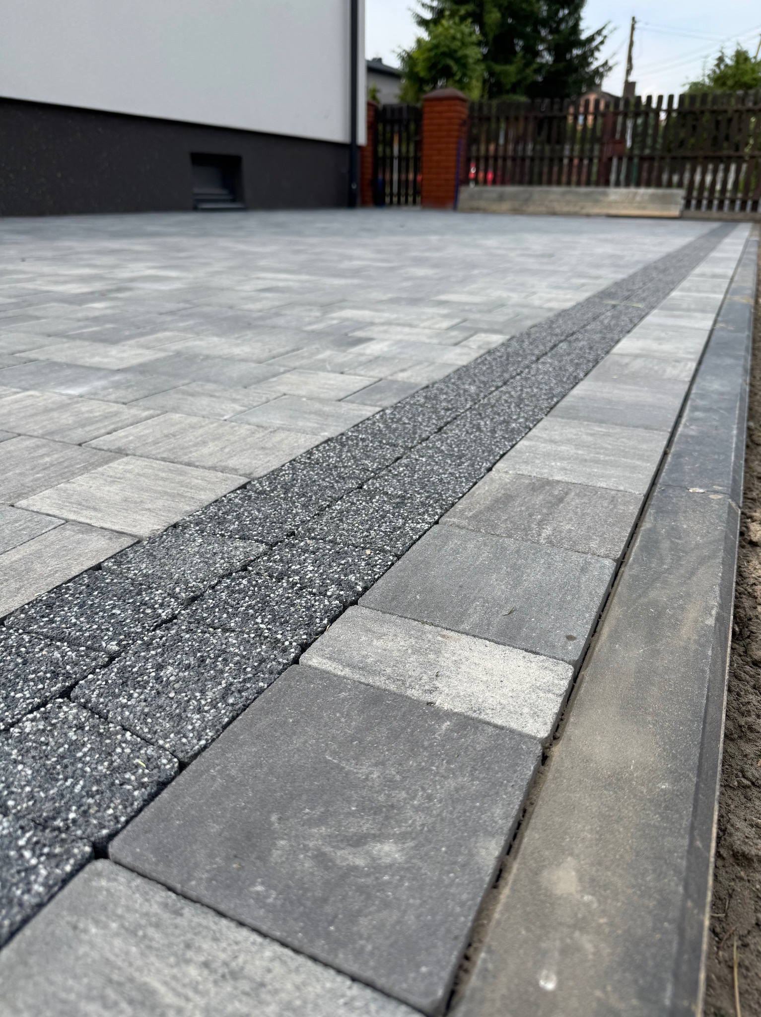 Ułożona szara kostka brukowa z ozdobnym pasem z drobnego grysu. Krawężnik oddziela nawierzchnię od ziemi. Widok na posesję z drewnianym płotem w tle.