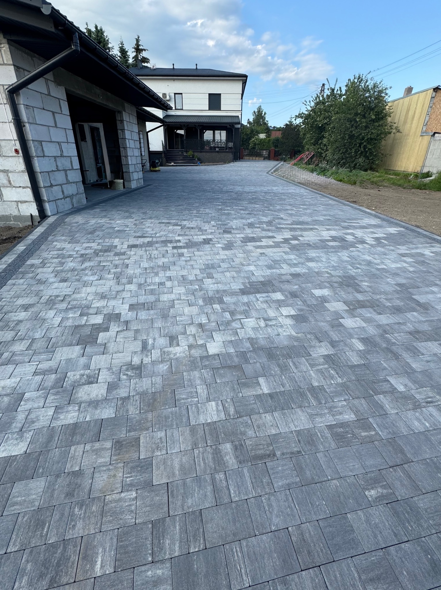 Nowo ułożona, długa aleja z szarej kostki brukowej prowadząca do domu z garażem w trakcie budowy, widok z perspektywy pieszego, błękitne niebo w tle.