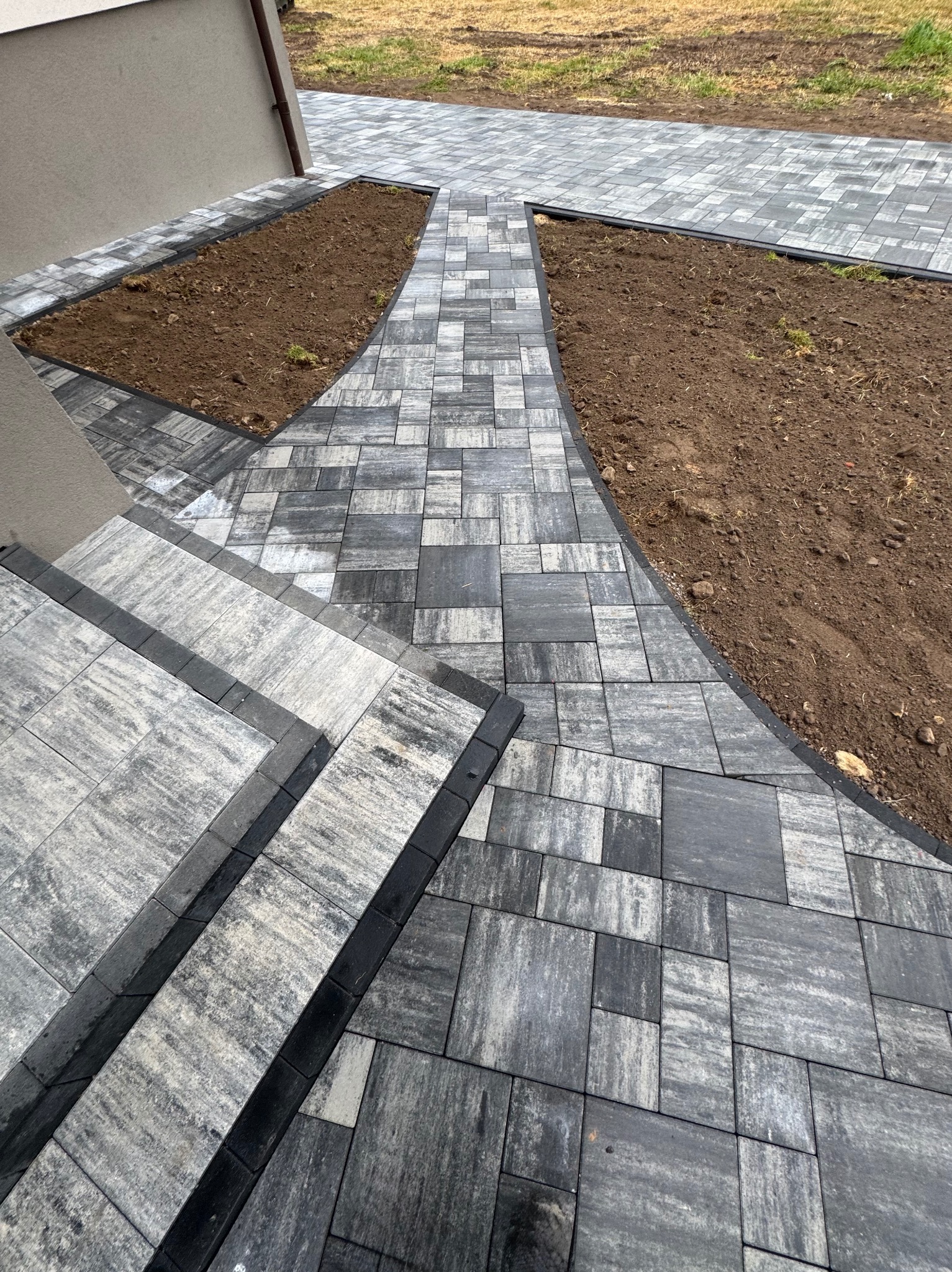 Ułożona kostka brukowa w odcieniach szarości prowadząca od schodów do ścieżki, z obrzeżami i rabatami ziemnymi po bokach. Widok z góry, świeżo po realizacji.