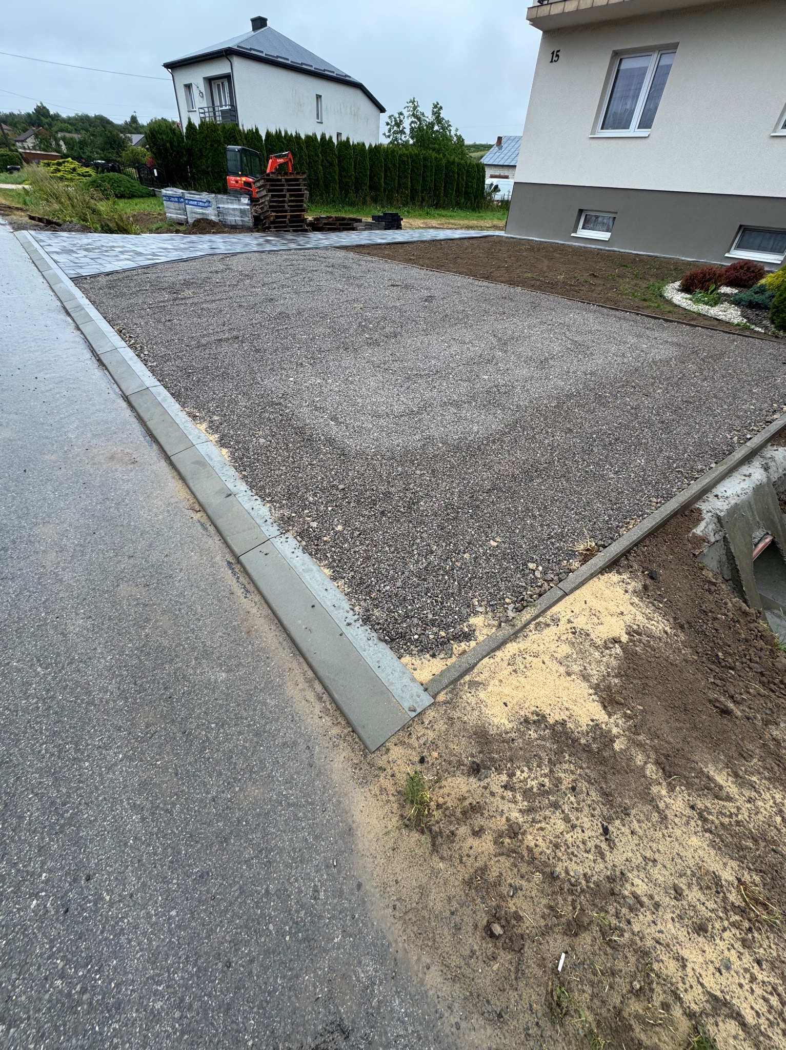 Przygotowany teren pod kostkę brukową z obrzeżami, zagęszczony żwir. W tle dom z numerem 15 i zaplecze z paletami kostki. Widok z poziomu gruntu.