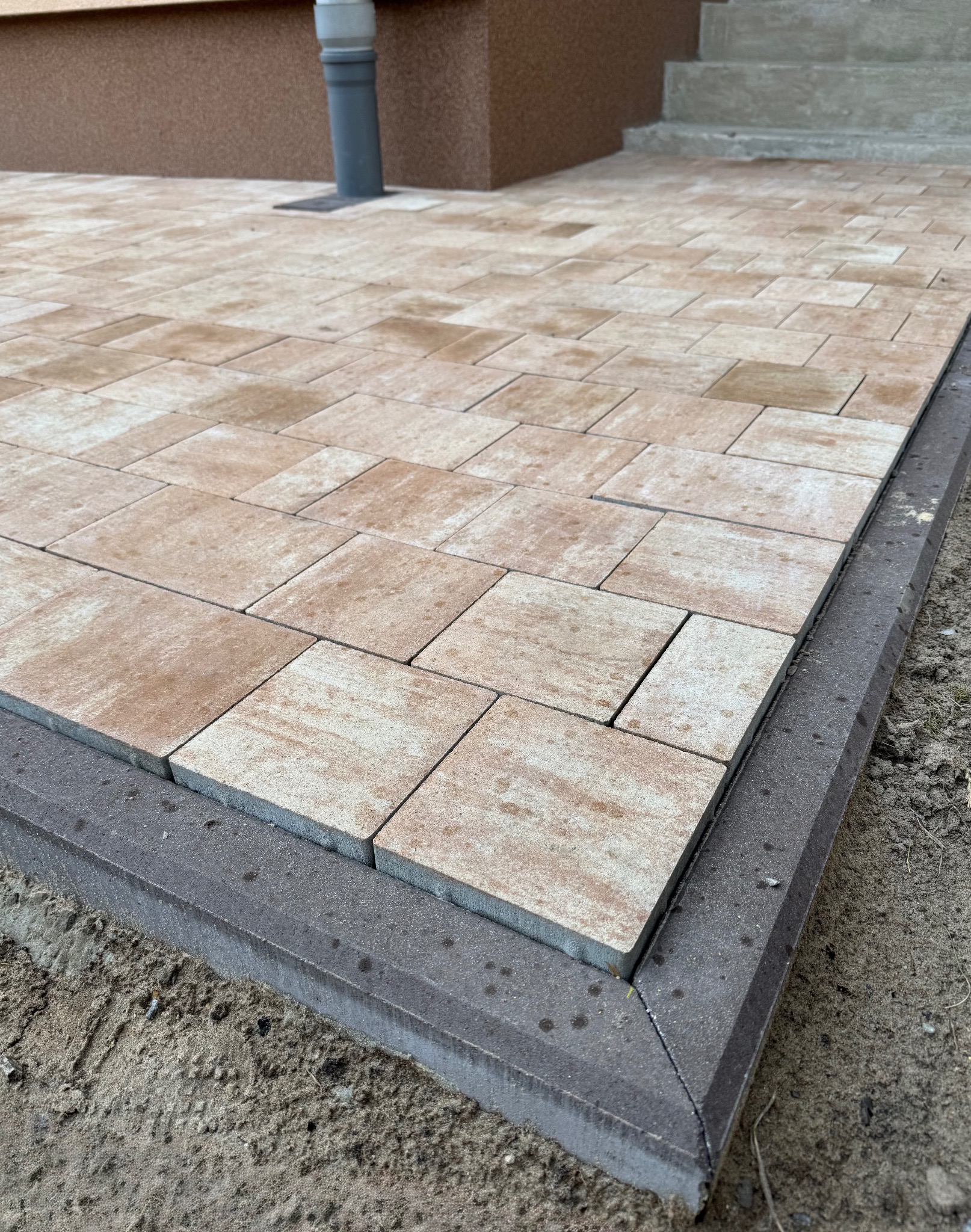 Beżowa kostka brukowa ułożona na posesji, obrzeża wykończone szarą kostką. Widok z góry, z bliska na fragment nawierzchni przy budynku i schodach.