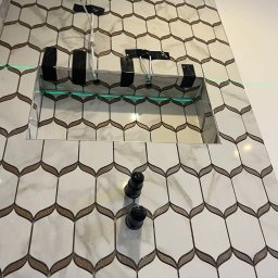 Łuk Drew Paweł Czekalinski - Ściana w łazience wyłożona białymi płytkami z marmurkowym wzorem i złotymi akcentami, widoczne przygotowane rury i wnęka w ścianie, wyznaczona linia lasera.