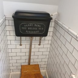 Łuk Drew Paweł Czekalinski - Retro toaleta z czarnym zbiornikiem 'Ferry Bank The Deluge No 2 Riverside' i drewnianą deską sedesową, białe kafelki na ścianach z czarną fugą.