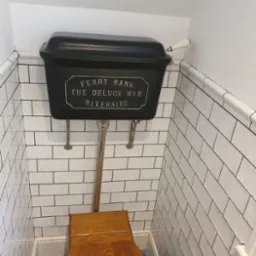 Retro toaleta z czarnym zbiornikiem 'Ferry Bank The Deluge No 2 Riverside' i drewnianą deską sedesową, białe kafelki na ścianach z czarną fugą.