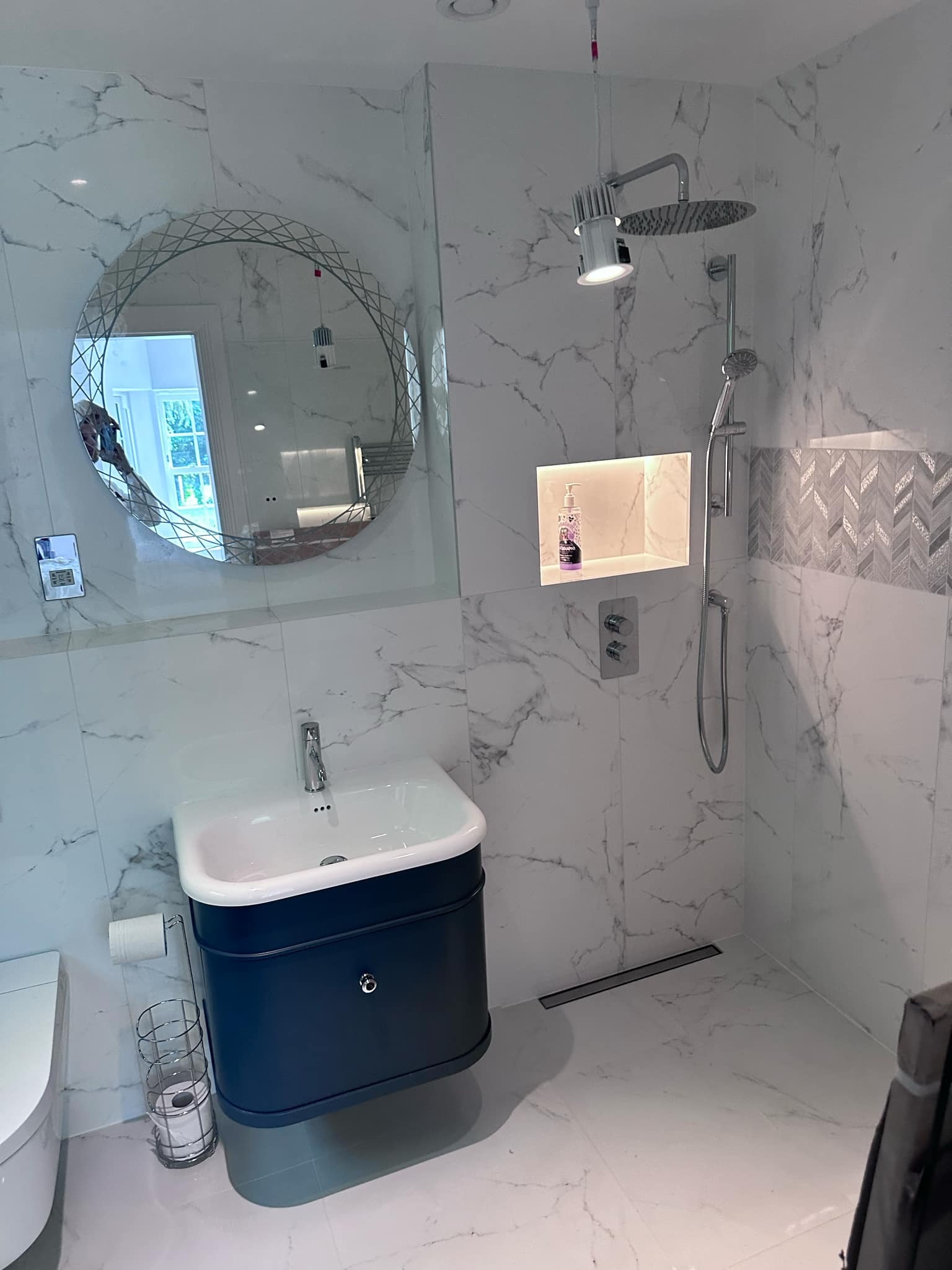 Nowoczesna łazienka z marmurowymi płytkami, okrągłym lustrem z dekoracyjną ramą, ciemnoniebieską szafką pod umywalką i deszczownicą z oświetleniem punktowym.