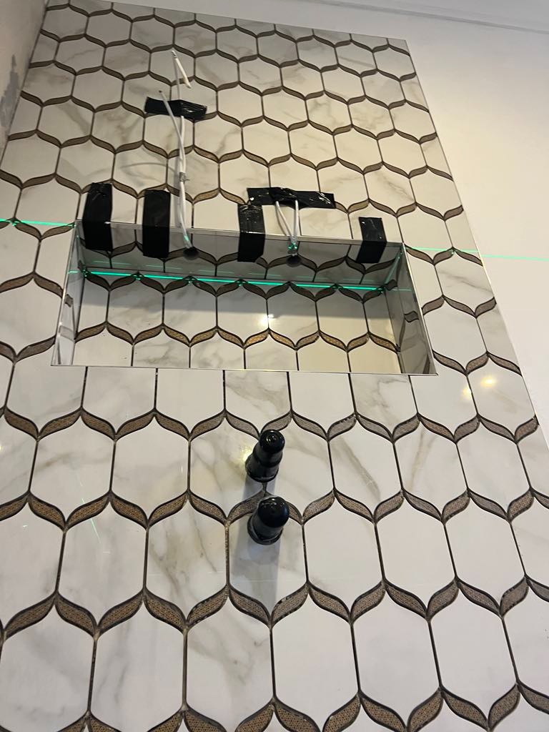 Ściana w łazience wyłożona białymi płytkami z marmurkowym wzorem i złotymi akcentami, widoczne przygotowane rury i wnęka w ścianie, wyznaczona linia lasera.