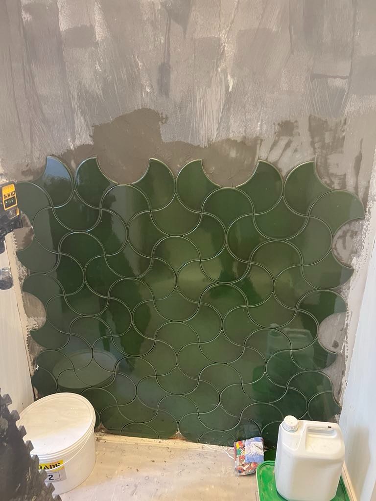 Układanie ciemnozielonych płytek w kształcie wachlarzy na ścianie, widoczne ślady zaprawy, narzędzia pracy w tle.