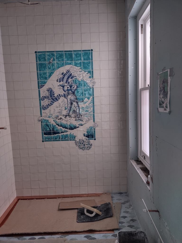 Ściana łazienki wyłożona białymi kwadratowymi płytkami z dekoracyjnym panelem z motywem fali i postaci, widoczny fragment okna, podłoga zabezpieczona kartonem, narzędzia glazurnicze