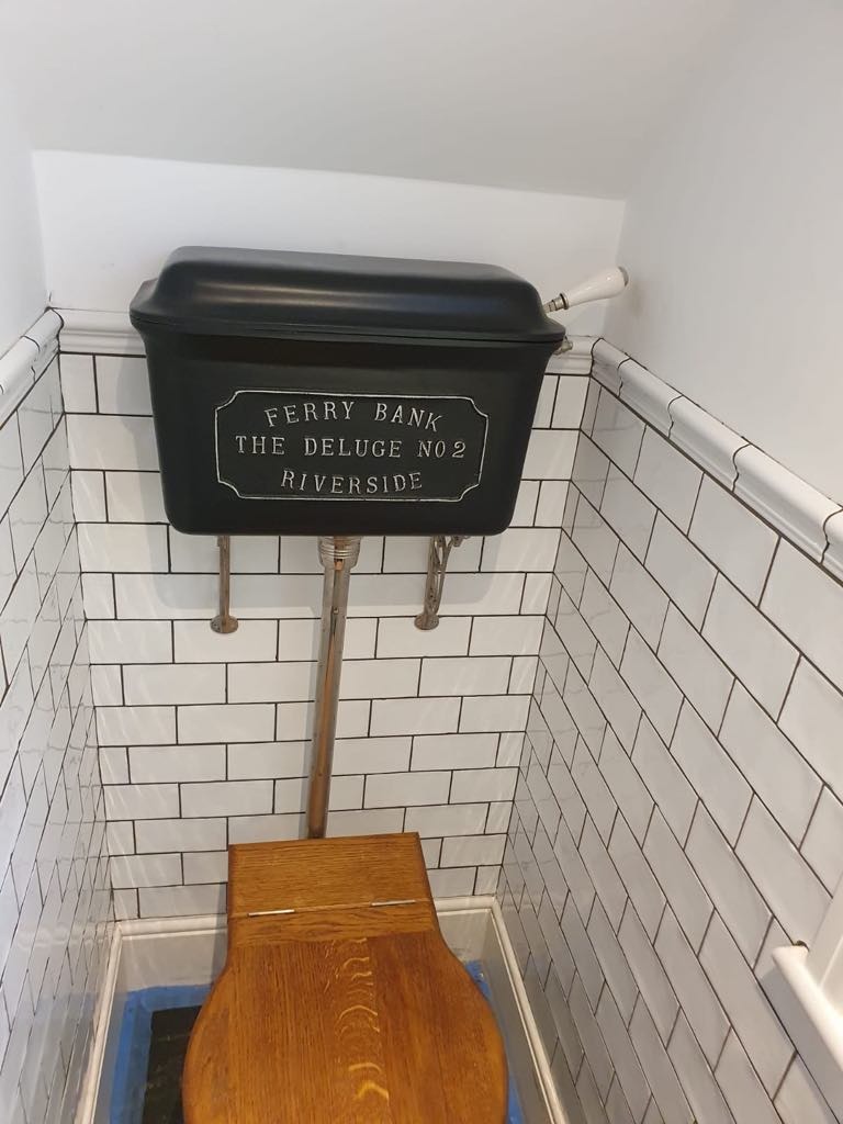 Retro toaleta z czarnym zbiornikiem 'Ferry Bank The Deluge No 2 Riverside' i drewnianą deską sedesową, białe kafelki na ścianach z czarną fugą.