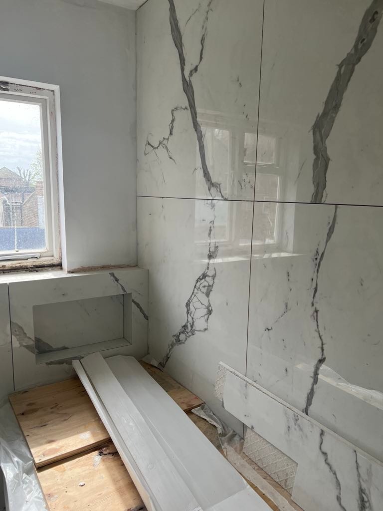 Łazienka w trakcie remontu: ściany wyłożone białymi płytami imitującymi marmur z szarymi żyłami, wnęka w ścianie, drewniane deski na podłodze, widoczne łączenia płyt.