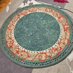 Okrągły, zielony dywan z wzorem w odcieniach czerwieni i beżu leżący na większym, wielobarwnym dywanie. Widoczny fragment parkietu i czarny kabel.