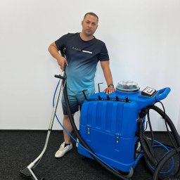 MasterClean - Mężczyzna opiera się o niebieski, profesjonalny odkurzacz do czyszczenia dywanów na tle białej ściany i ciemnej wykładziny. Widoczny wąż i dysza odkurzacza.