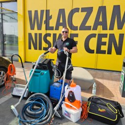 MasterClean - Profesjonalny zestaw do czyszczenia tapicerki i dywanów z operatorem na tle żółtej ściany z napisem 'Włączam niskie ceny'. Widoczne akcesoria i środki czystości.