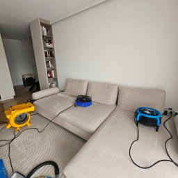MasterClean - Beżowa sofa modułowa z wentylatorami i sprzętem czyszczącym na wzorzystym dywanie. W tle regał z książkami i wejście do innego pomieszczenia.