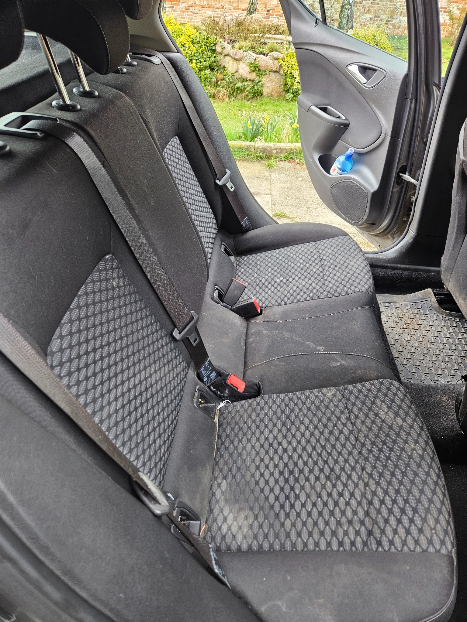 Brudna tylna kanapa samochodu z widocznymi plamami i zabrudzeniami na tapicerce oraz dywaniku podłogowym, z otwartymi drzwiami i butelką wody w schowku.