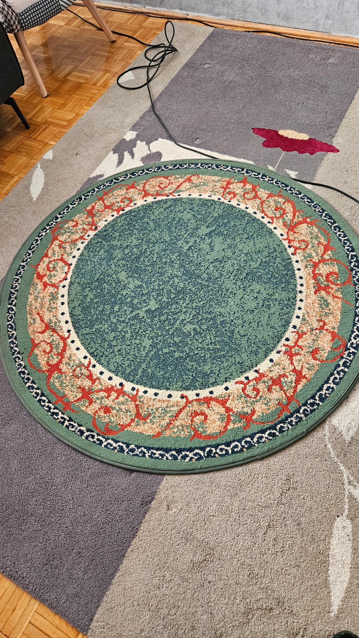 Okrągły, zielony dywan z wzorem w odcieniach czerwieni i beżu leżący na większym, wielobarwnym dywanie. Widoczny fragment parkietu i czarny kabel.