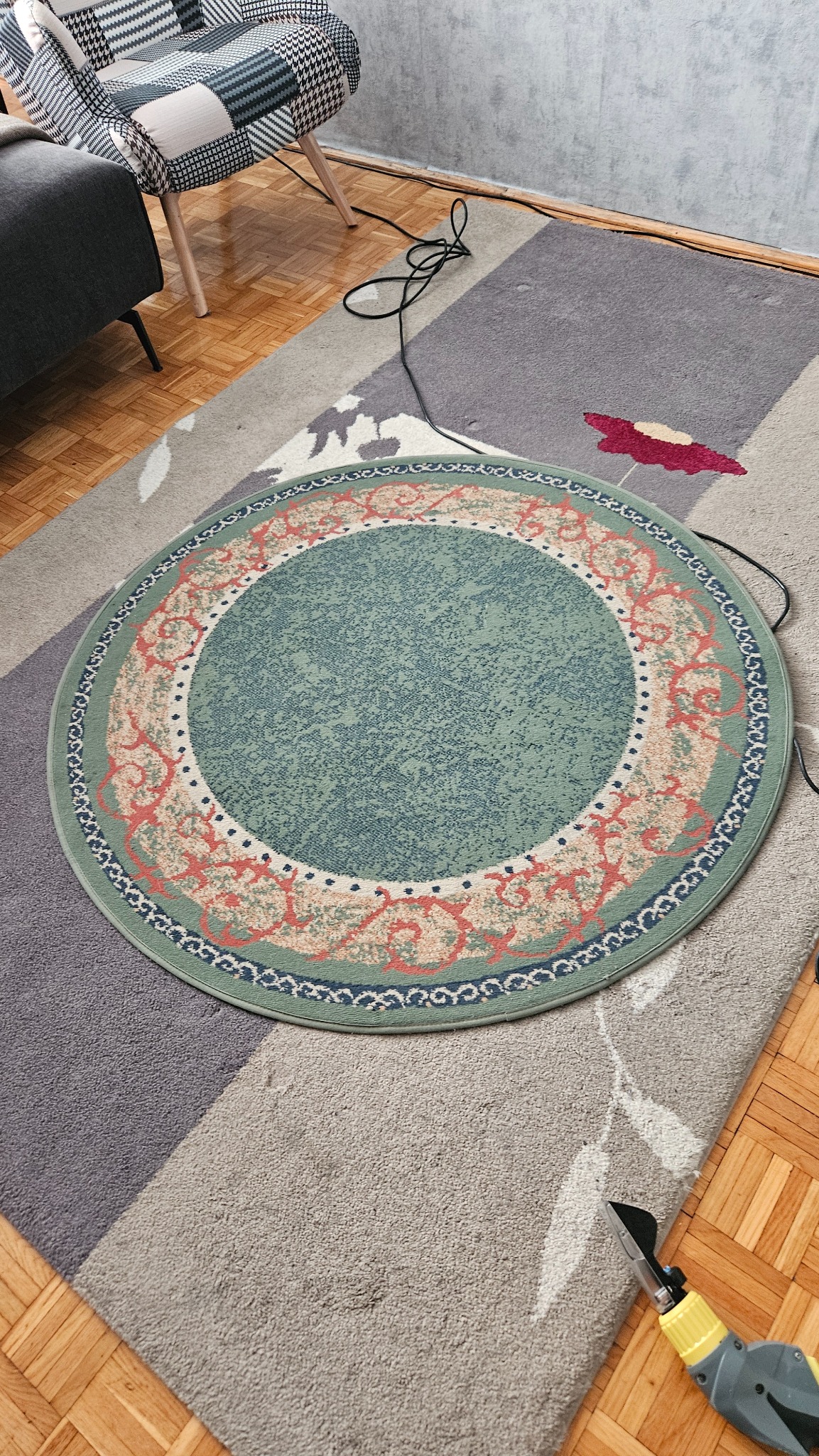 Okrągły, wzorzysty dywan na podłodze obok krzesła i sofy. Na dywanie leży narzędzie z żółtą rączką.