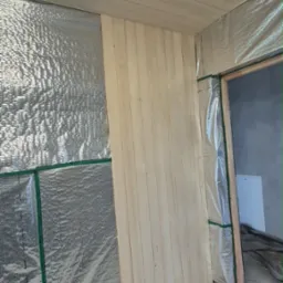 Wnętrze w trakcie wykańczania: ściana z izolacją pokryta folią aluminiową i częściowo obłożona jasnym drewnem, widoczny sufit z paneli i fragment podłogi.