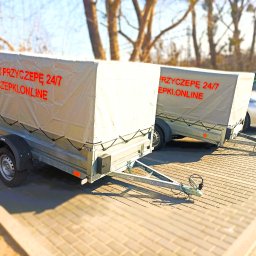 Przyczepki.Online - Dwie przyczepy towarowe z plandekami z napisem 'Wynajmij przyczepę 24/7 przyczepki.online' stojące na brukowanym placu, w tle drzewa i zaparkowane samochody.