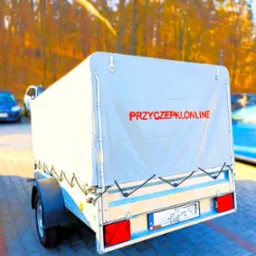 Przyczepka samochodowa z plandeką, na której widnieje czerwony napis PRZYCZEPKI.ONLINE, zaparkowana na brukowanym placu z drzewami w tle.