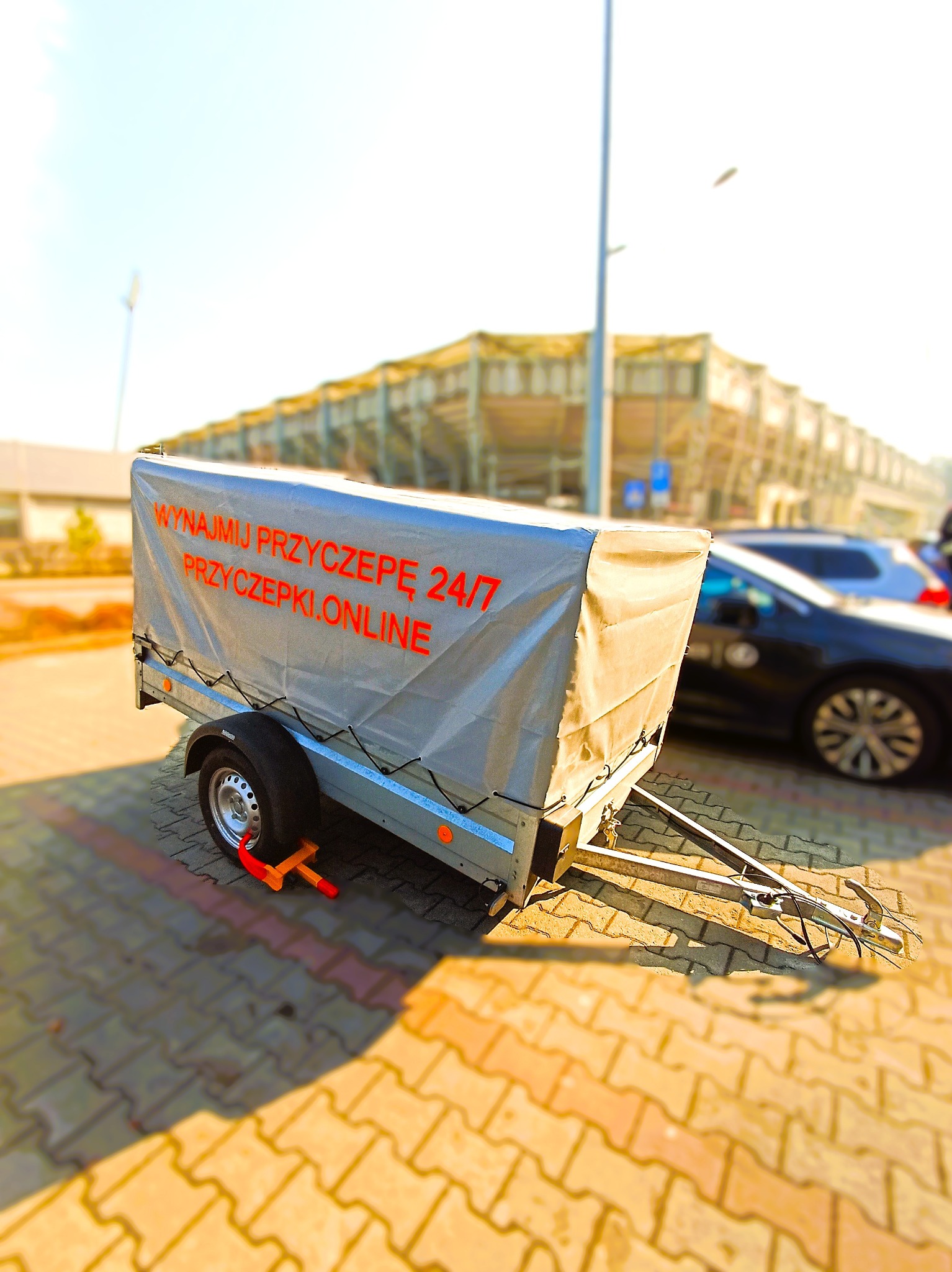 Przyczepa z plandeką z napisem 'Wynajmij przyczepę 24/7 przyczepki.online' zaparkowana na brukowanym placu, zabezpieczona pomarańczową blokadą na koło, w tle budynek.