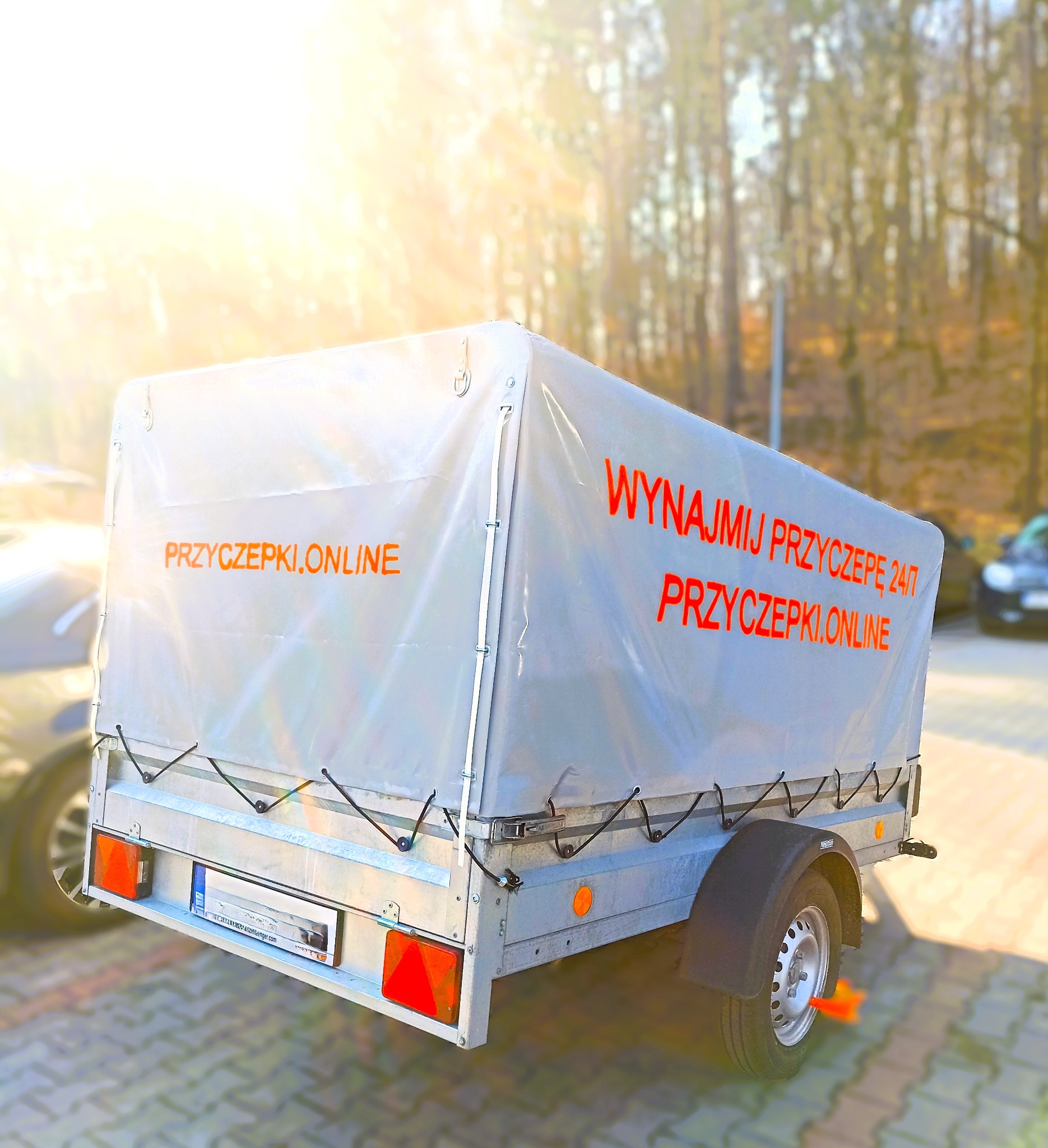 Przyczepa z plandeką, na której widnieje napis 'Wynajmij przyczepę 24/7 Przyczepki.online', zaparkowana na brukowanym placu, w tle drzewa i samochód, oświetlona promieniami słońca.