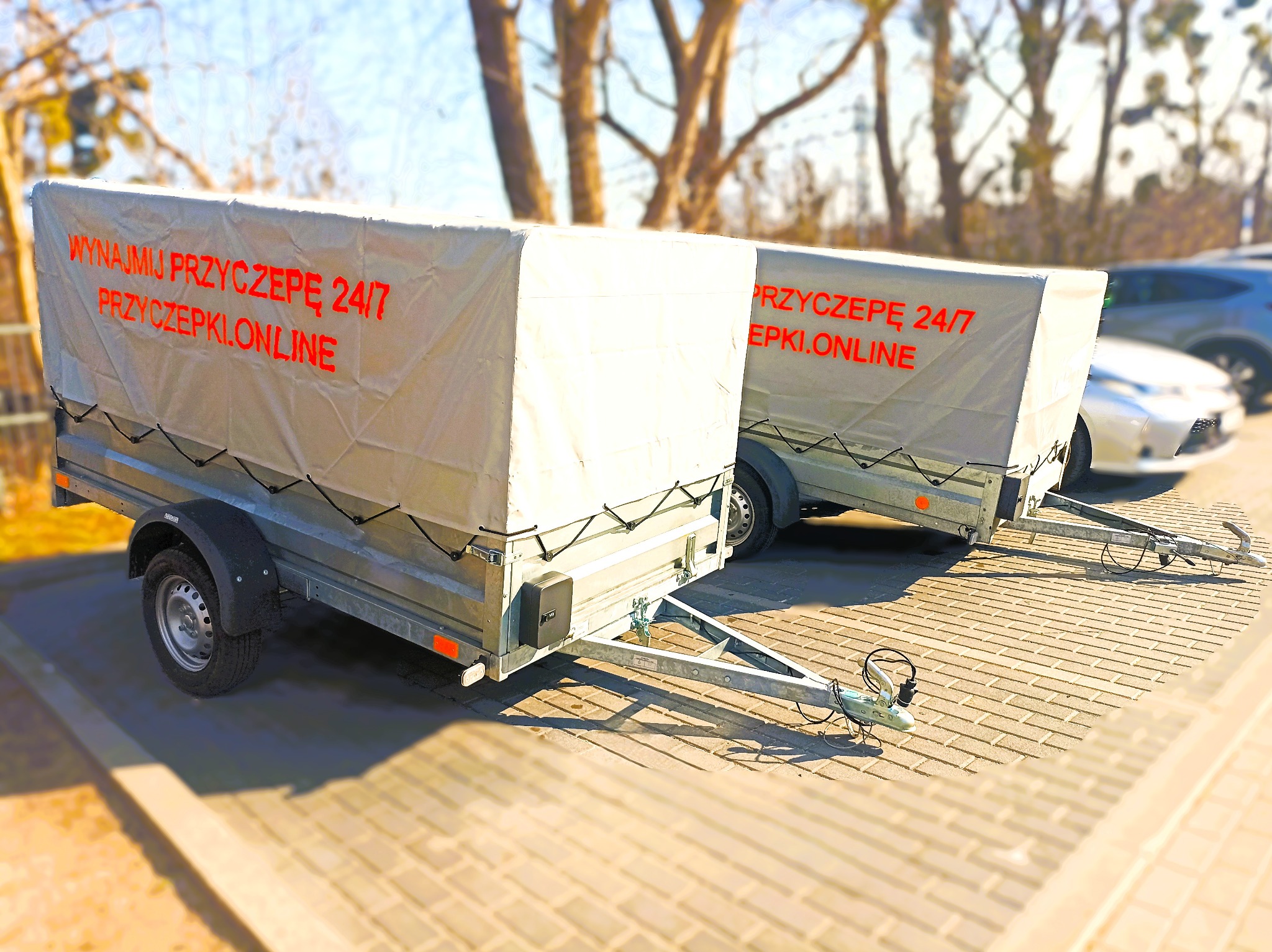 Dwie przyczepy towarowe z plandekami z napisem 'Wynajmij przyczepę 24/7 przyczepki.online' stojące na brukowanym placu, w tle drzewa i zaparkowane samochody.