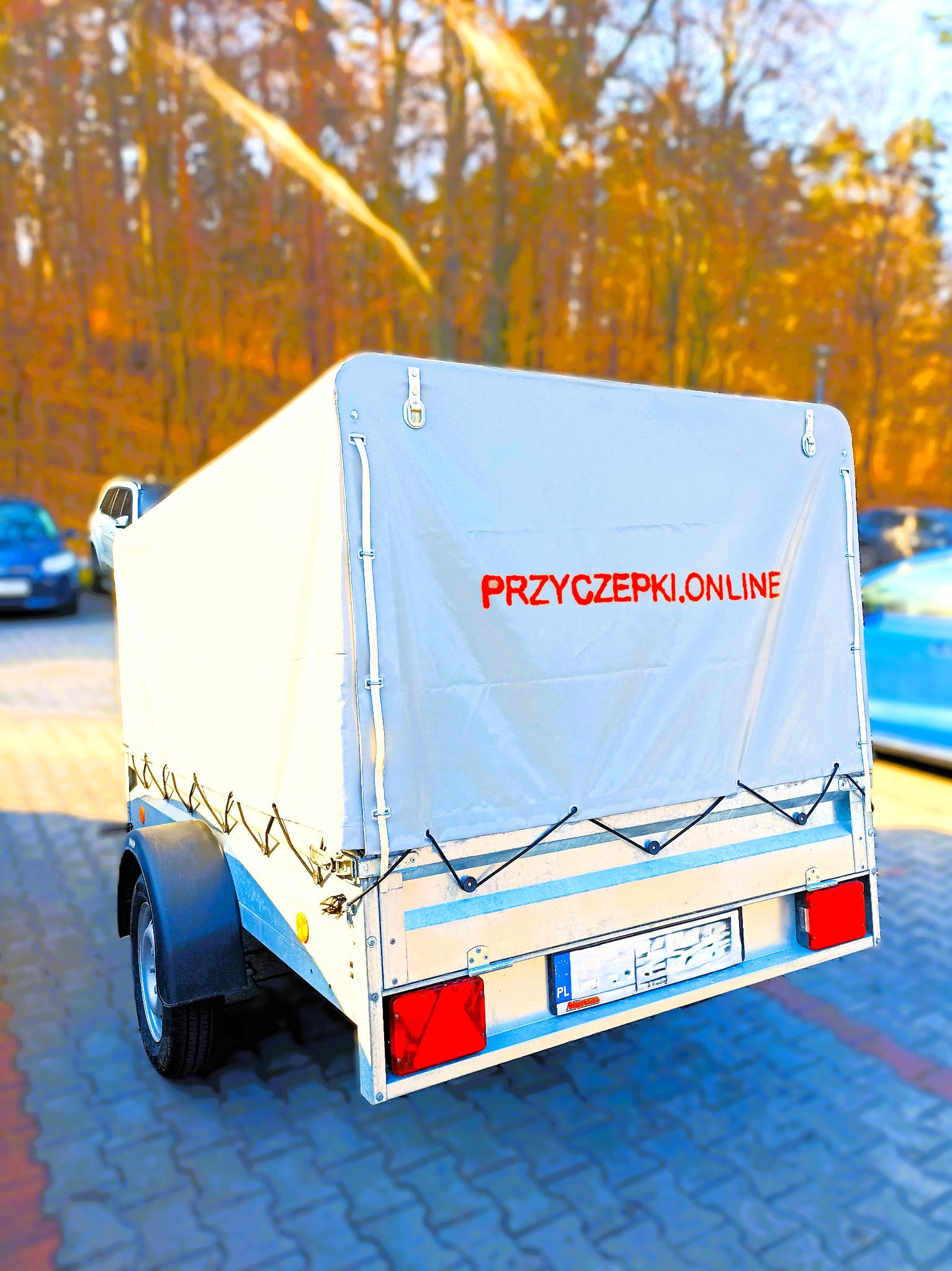Przyczepka samochodowa z plandeką, na której widnieje czerwony napis PRZYCZEPKI.ONLINE, zaparkowana na brukowanym placu z drzewami w tle.