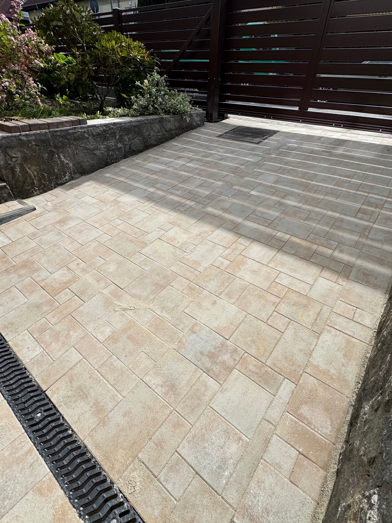 Beżowa kostka brukowa ułożona na podjeździe, z widoczną linią drenażową i bramą w tle, w słoneczny dzień. Różne rozmiary kostek tworzą unikalny wzór.