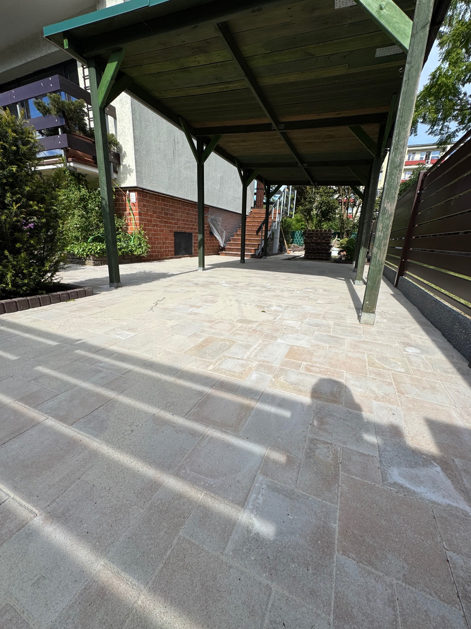 Jasne kostki brukowe pod drewnianą wiatą samochodową, z widocznym fragmentem domu i zielenią w tle. Cień rzucany przez konstrukcję tworzy geometryczny wzór.