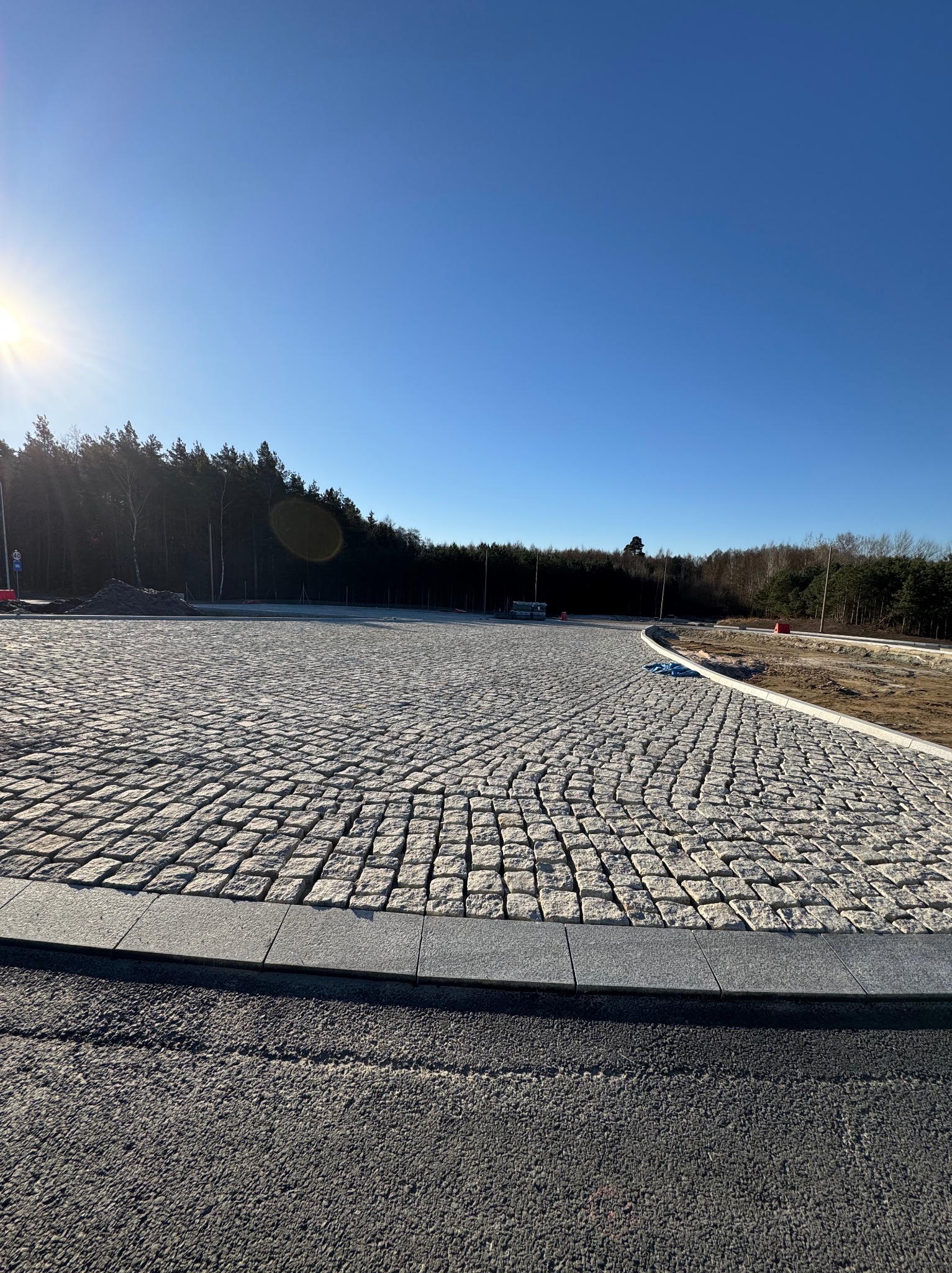 Rozległy plac wyłożony kostką granitową, otoczony obrzeżami. W tle widoczny las i błękitne niebo z promieniami słońca. Nowoczesne i estetyczne wykończenie.