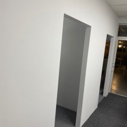 art.room - Biała ściana z dwoma otworami drzwiowymi, świeżo pomalowana, widok z dołu, szara wykładzina podłogowa, jasne oświetlenie, wnętrze biura lub mieszkania.