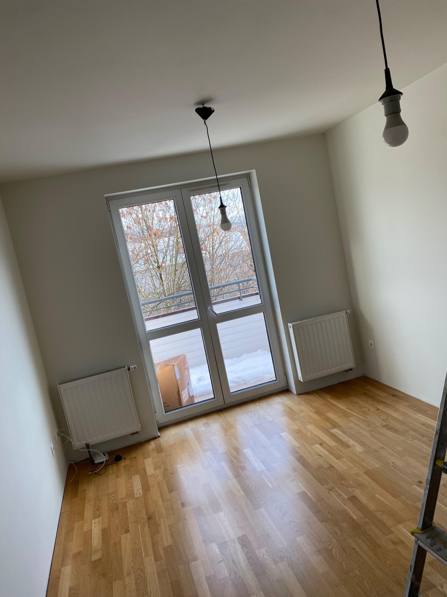 Pusty pokój po remoncie z parkietem, białymi ścianami, grzejnikami i oknem balkonowym z widokiem na zaśnieżony balkon. Wiszące żarówki i fragment drabiny.
