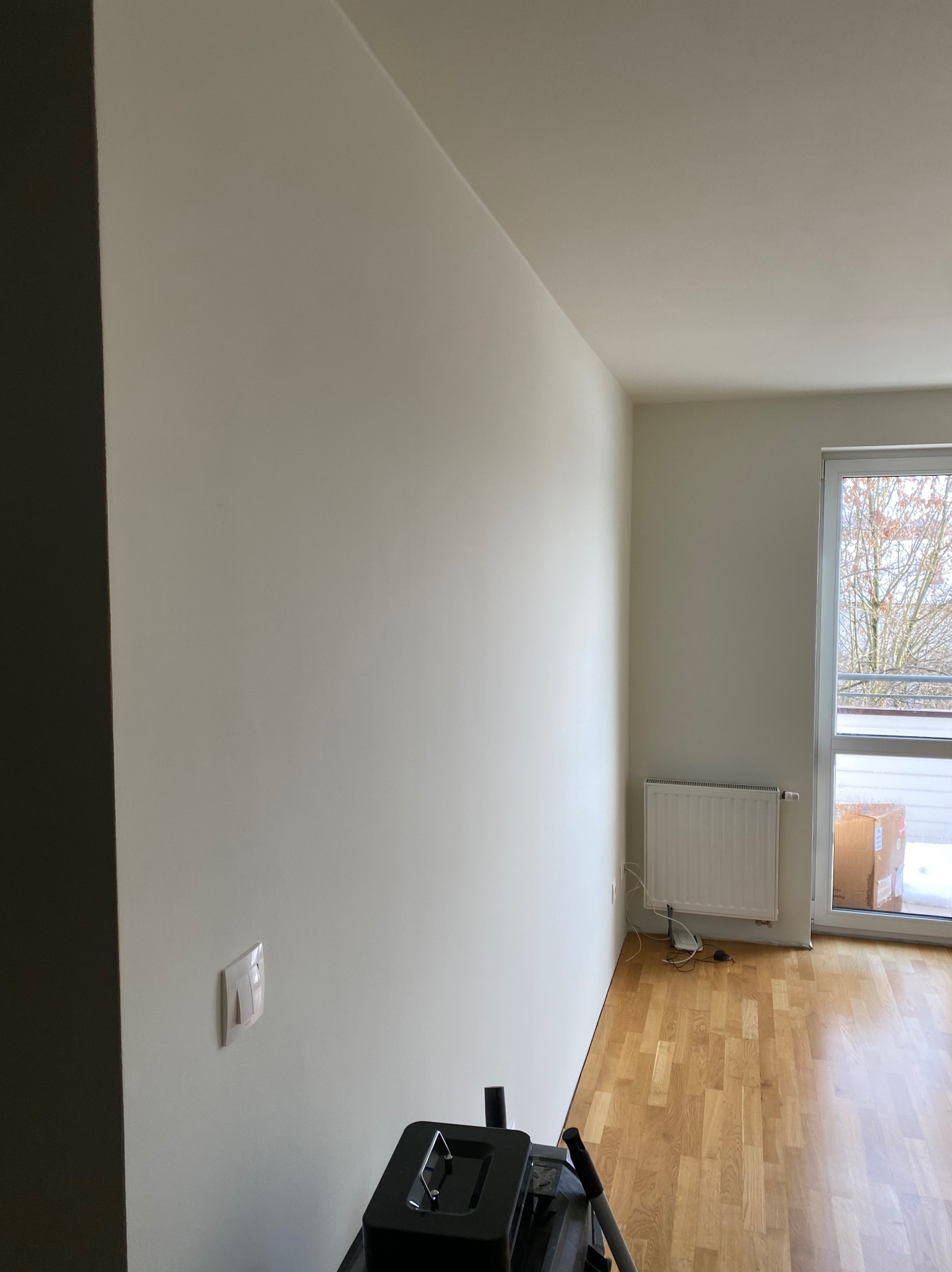 Jasne, świeżo pomalowane wnętrze z parkietem i grzejnikiem pod oknem. Widoczny fragment sprzętu malarskiego w dolnej części kadru.