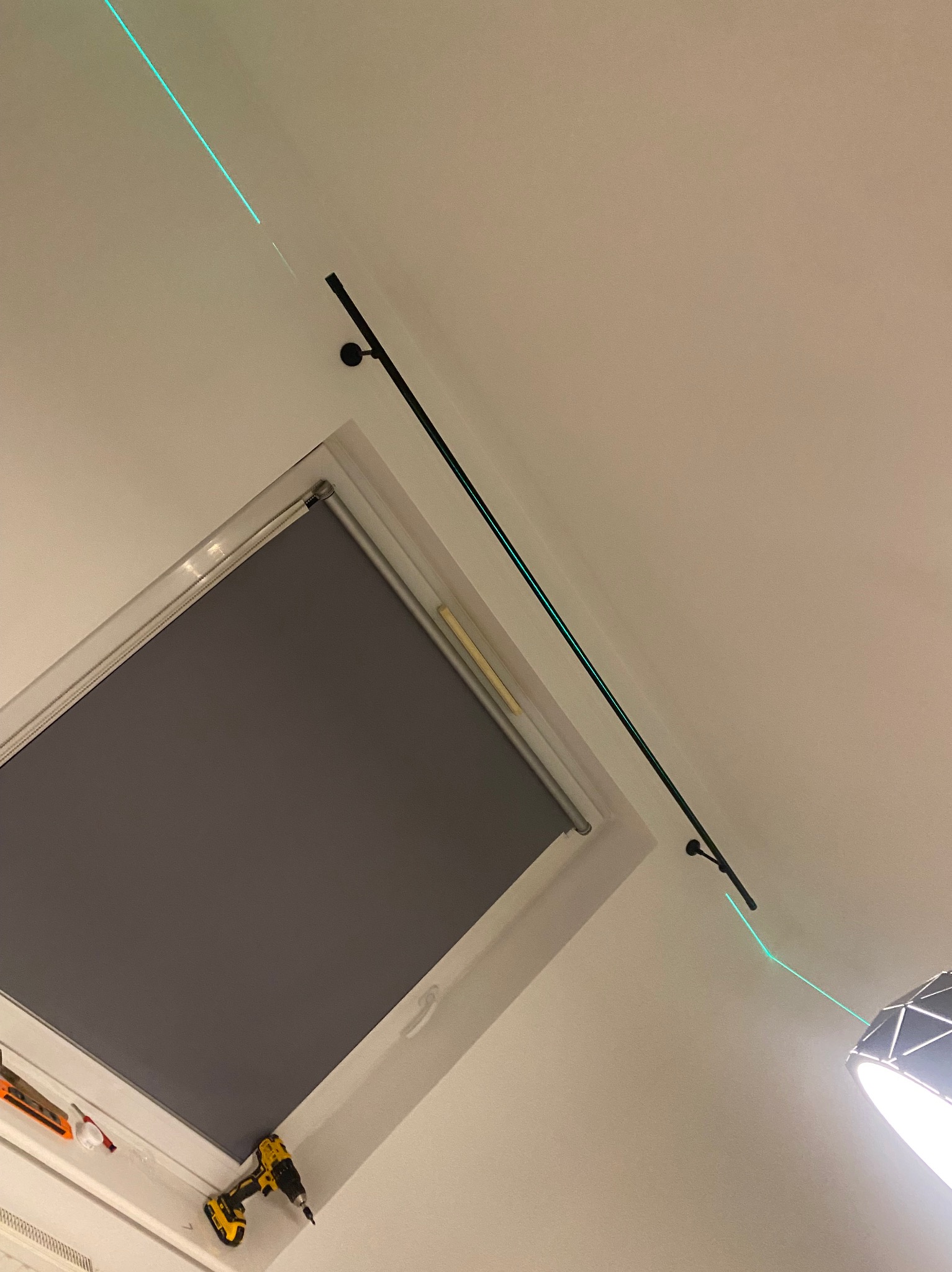 Montaż karnisza na suficie pod skosem, widoczny laser wyznaczający linię, okno dachowe z roletą i wkrętarka. Nietypowy kąt ujęcia.