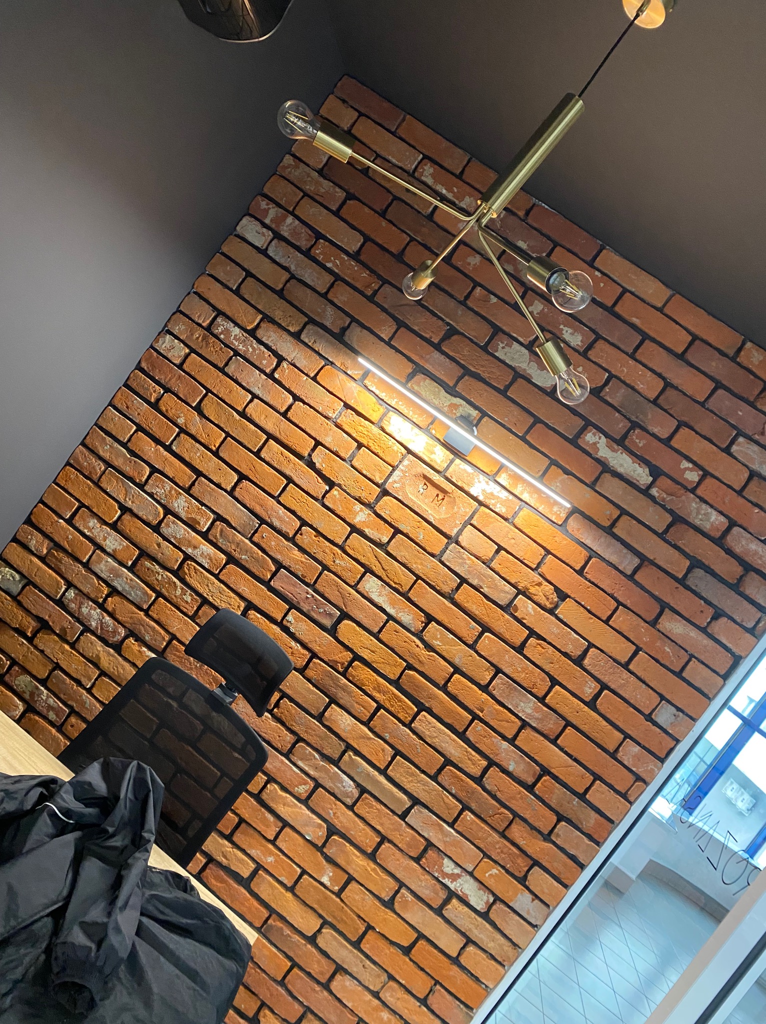 Ceglana ściana z oświetleniem LED i designerską lampą sufitową w nowoczesnym biurze. Widoczny fragment biurka i krzesła.