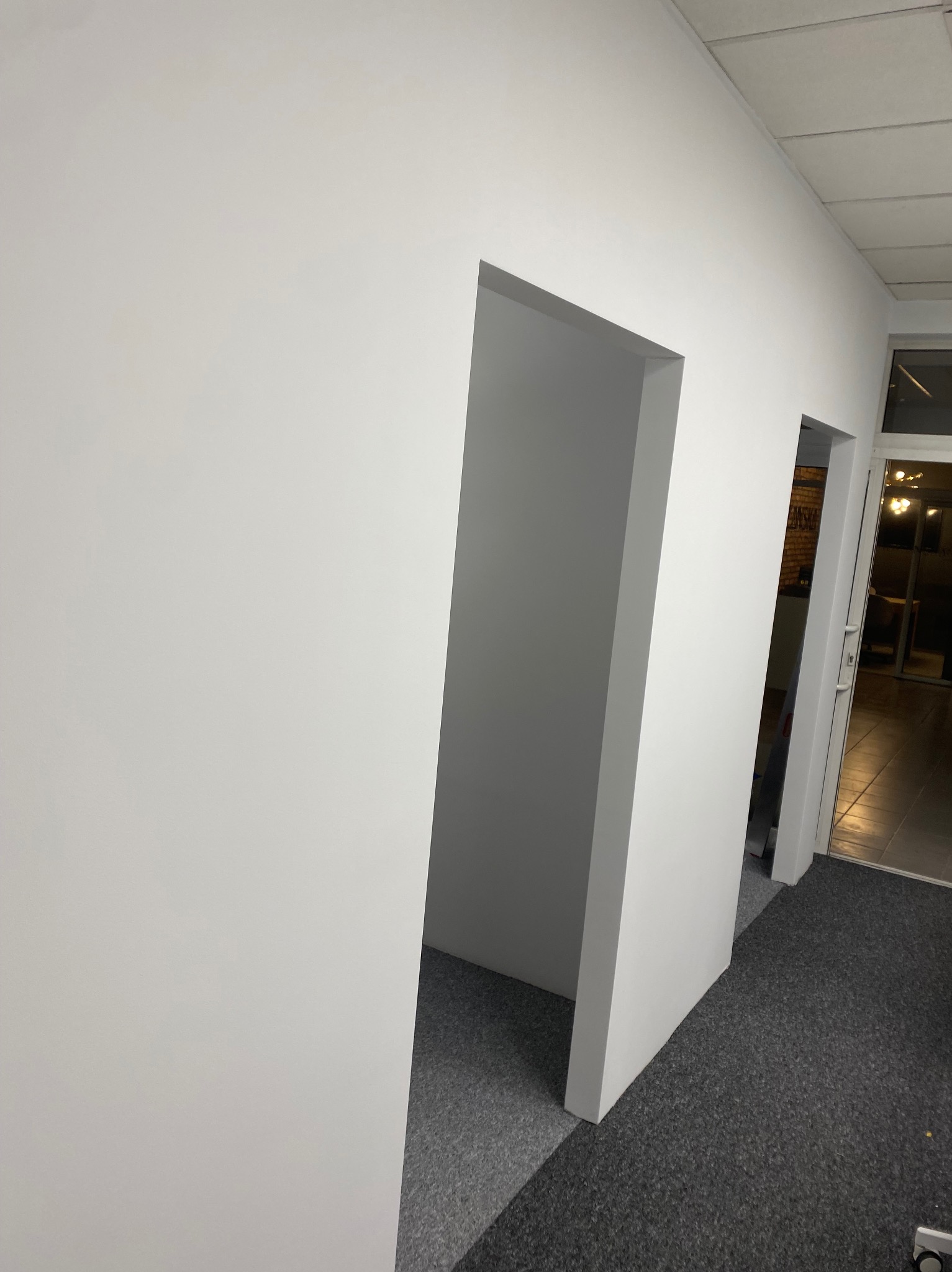 Biała ściana z dwoma otworami drzwiowymi, świeżo pomalowana, widok z dołu, szara wykładzina podłogowa, jasne oświetlenie, wnętrze biura lub mieszkania.