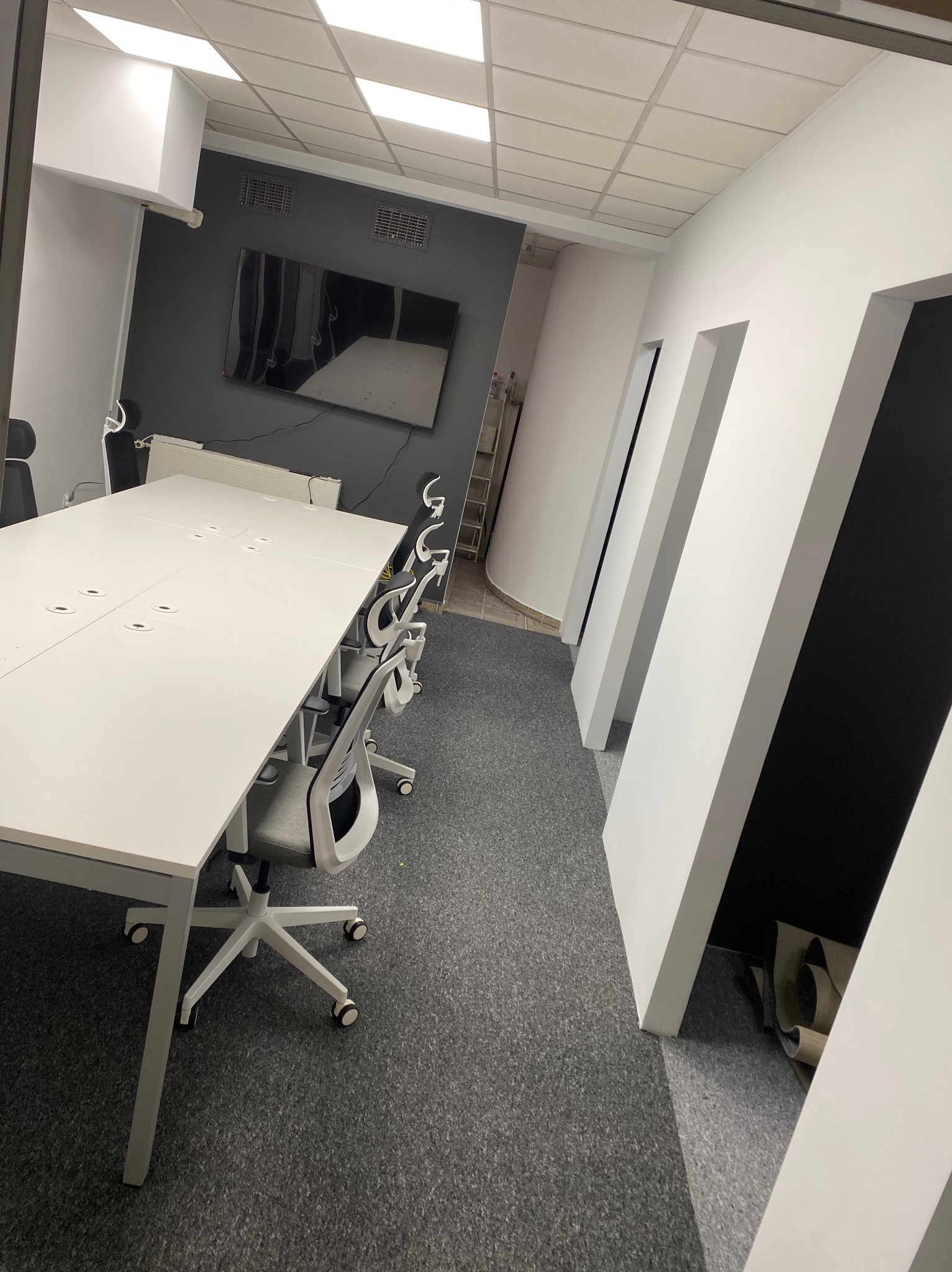 Nowoczesne biuro po remoncie: biały stół konferencyjny, szare krzesła obrotowe, ciemnoszara ściana z telewizorem, białe ściany z czarnymi wnękami. Wykończenie wnętrz biura.