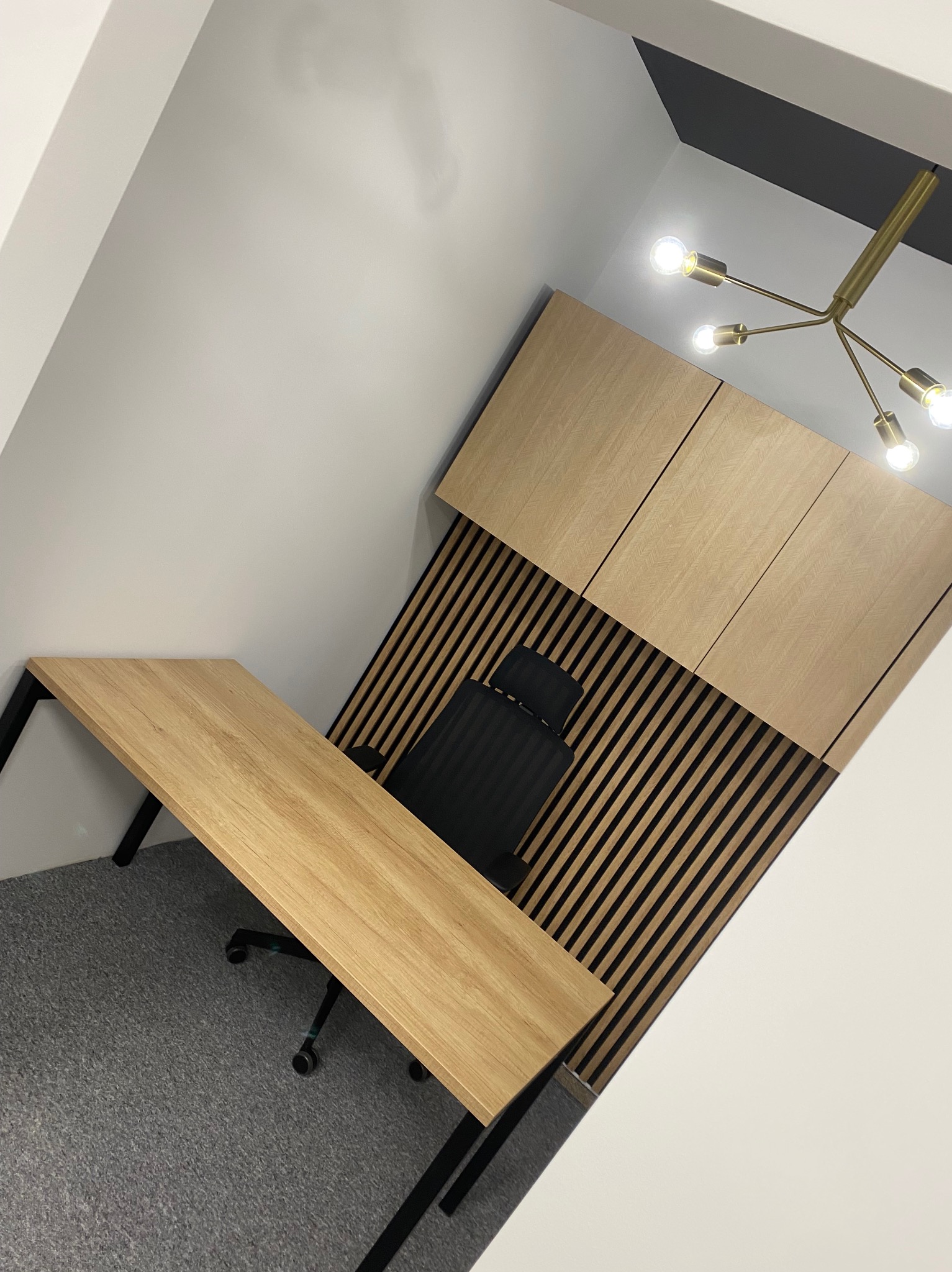 Nowoczesne biuro: drewniane biurko z czarnymi nogami, krzesło biurowe, lamelowa ściana i szafki w odcieniach drewna, oświetlone złotą lampą sufitową.