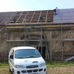 Atila dekarz - Renowacja dachu starego budynku gospodarczego: ekipa monterów na rusztowaniu układa metalowe arkusze na drewnianej konstrukcji, część dachu pokryta starym materiałem, na pierwszym planie biały...