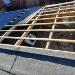 Montaż drewnianej konstrukcji dachu z widoczną częścią pokrycia z blachy trapezowej oraz pracownikiem na dachu budynku.