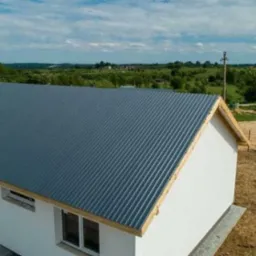 Nowoczesny dom jednorodzinny z szarą blachą trapezową na dachu, widok z góry, surowy stan wykończenia terenu wokół budynku.