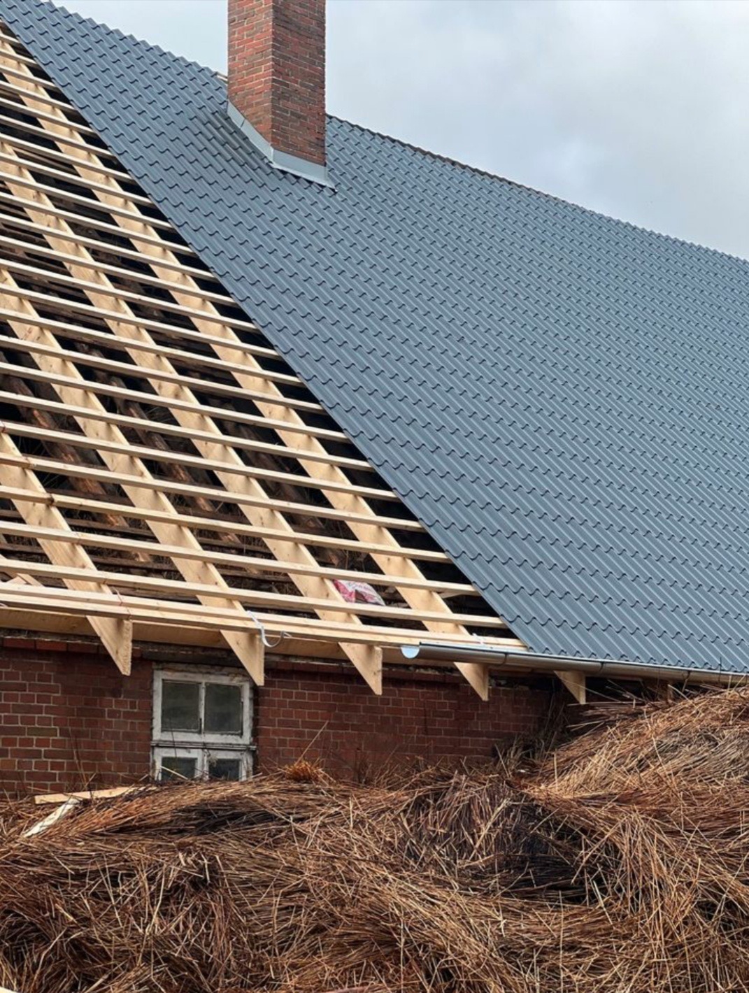Częściowo pokryty dach z szarej blachy falistej, widoczna drewniana konstrukcja więźby dachowej i ceglany komin, sterta suchych roślin u podnóża budynku.