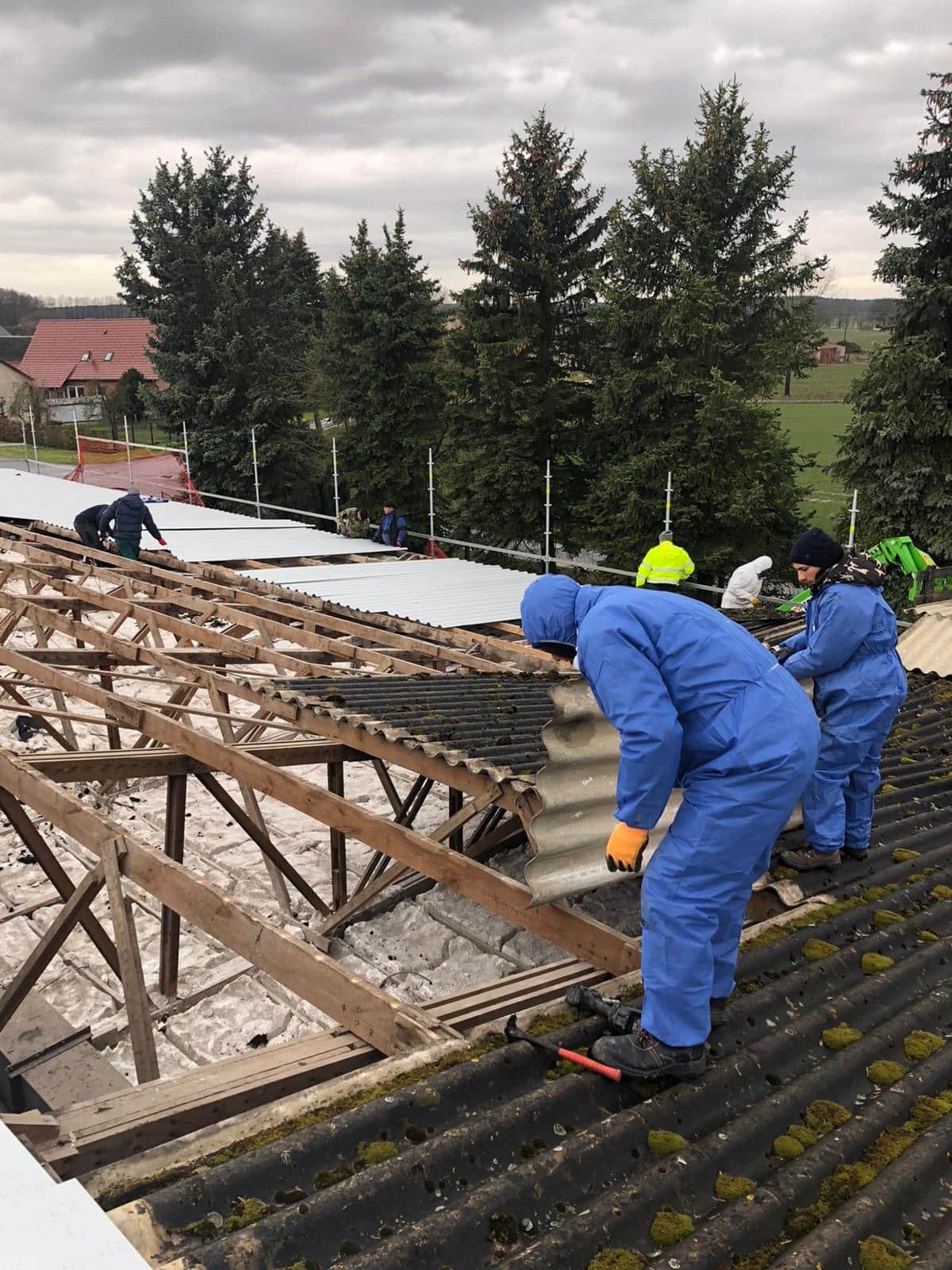 Pracownicy w niebieskich kombinezonach demontują stare, pokryte mchem płyty eternitowe z dachu, odsłaniając drewnianą konstrukcję więźby i białą izolację. W tle widoczne nowe, jasne arkusze...