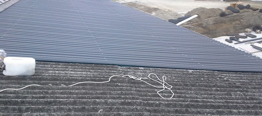 Stara, pokryta mchem dachówka sąsiaduje z nową, szarą blachą trapezową, na dachu leży biały pojemnik i zwinięty sznur, w tle hałda ziemi i materiały budowlane.