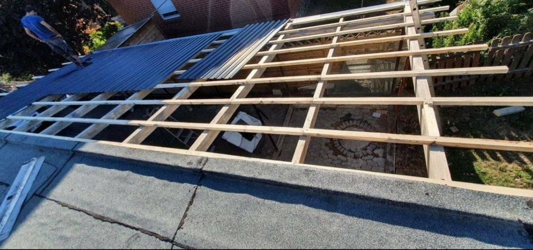 Montaż drewnianej konstrukcji dachu z widoczną częścią pokrycia z blachy trapezowej oraz pracownikiem na dachu budynku.