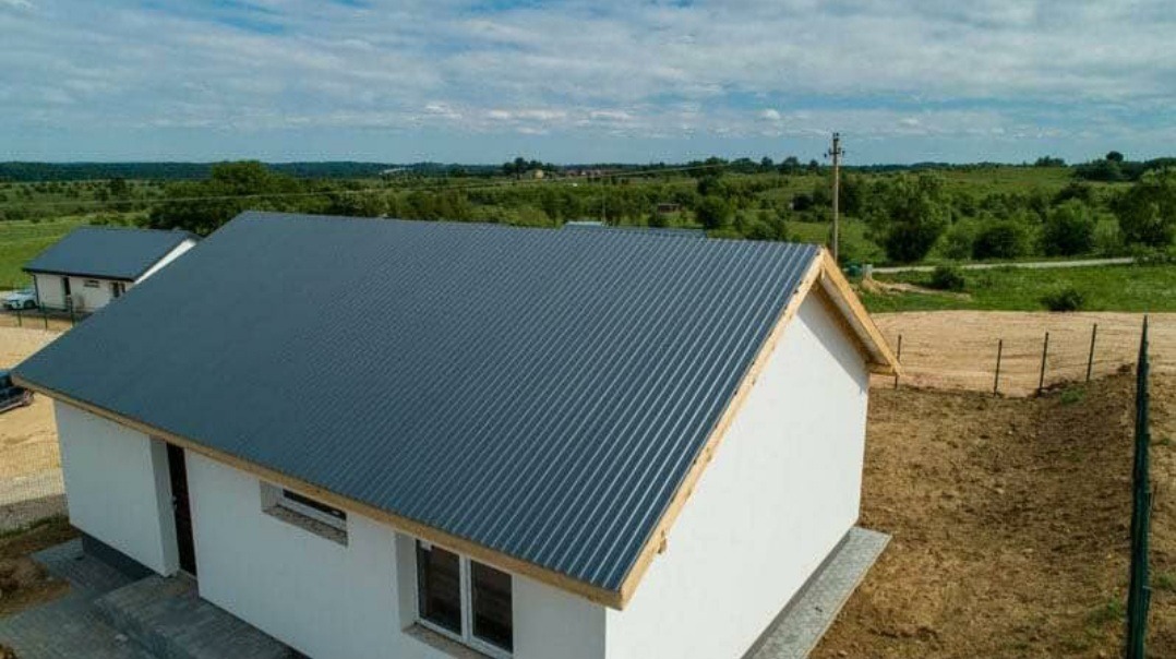 Nowoczesny dom jednorodzinny z szarą blachą trapezową na dachu, widok z góry, surowy stan wykończenia terenu wokół budynku.